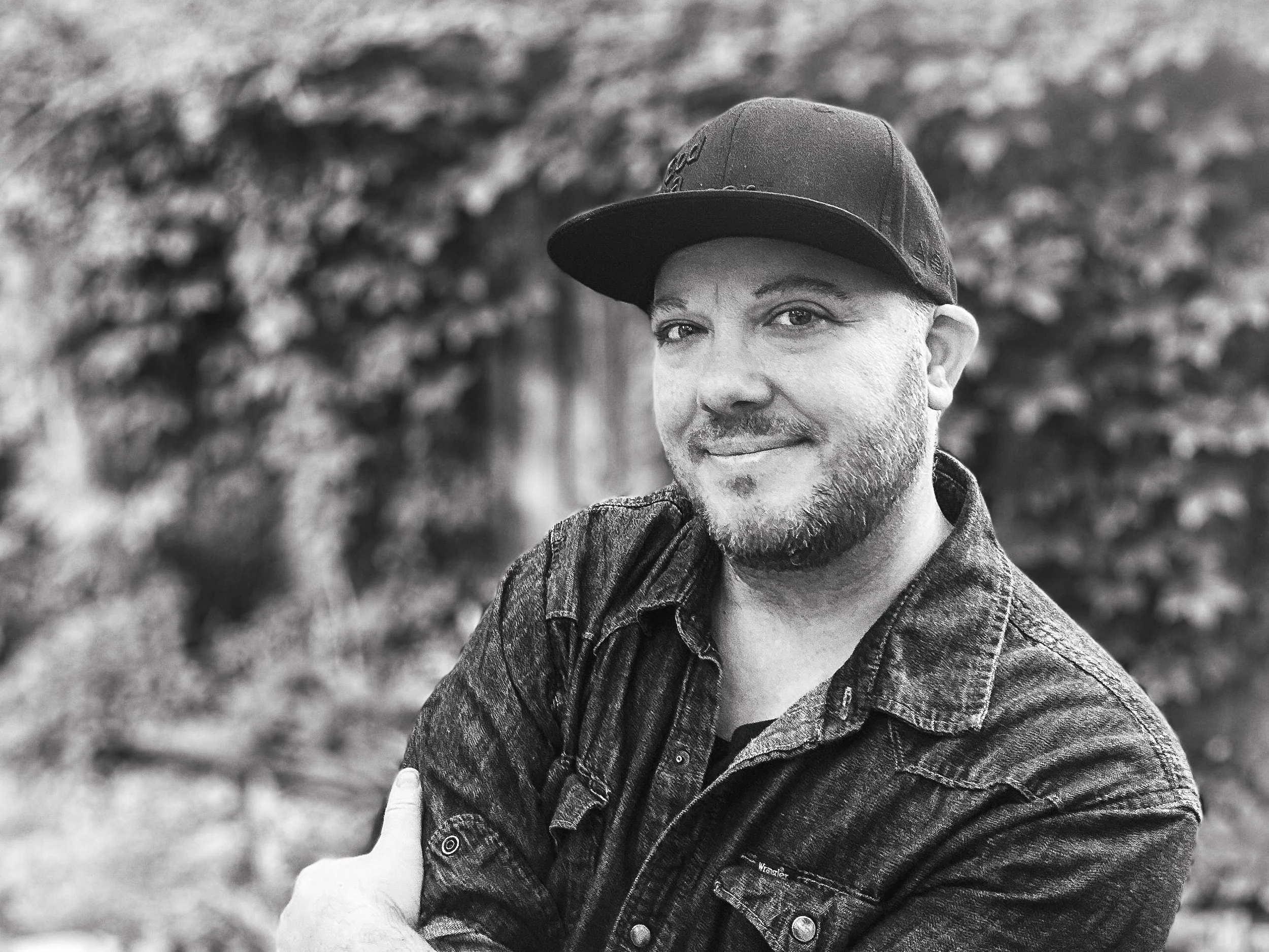 A black and white photo of a man wearing a baseball cap and denim jacket, smiling outdoors with a blurred natural background.