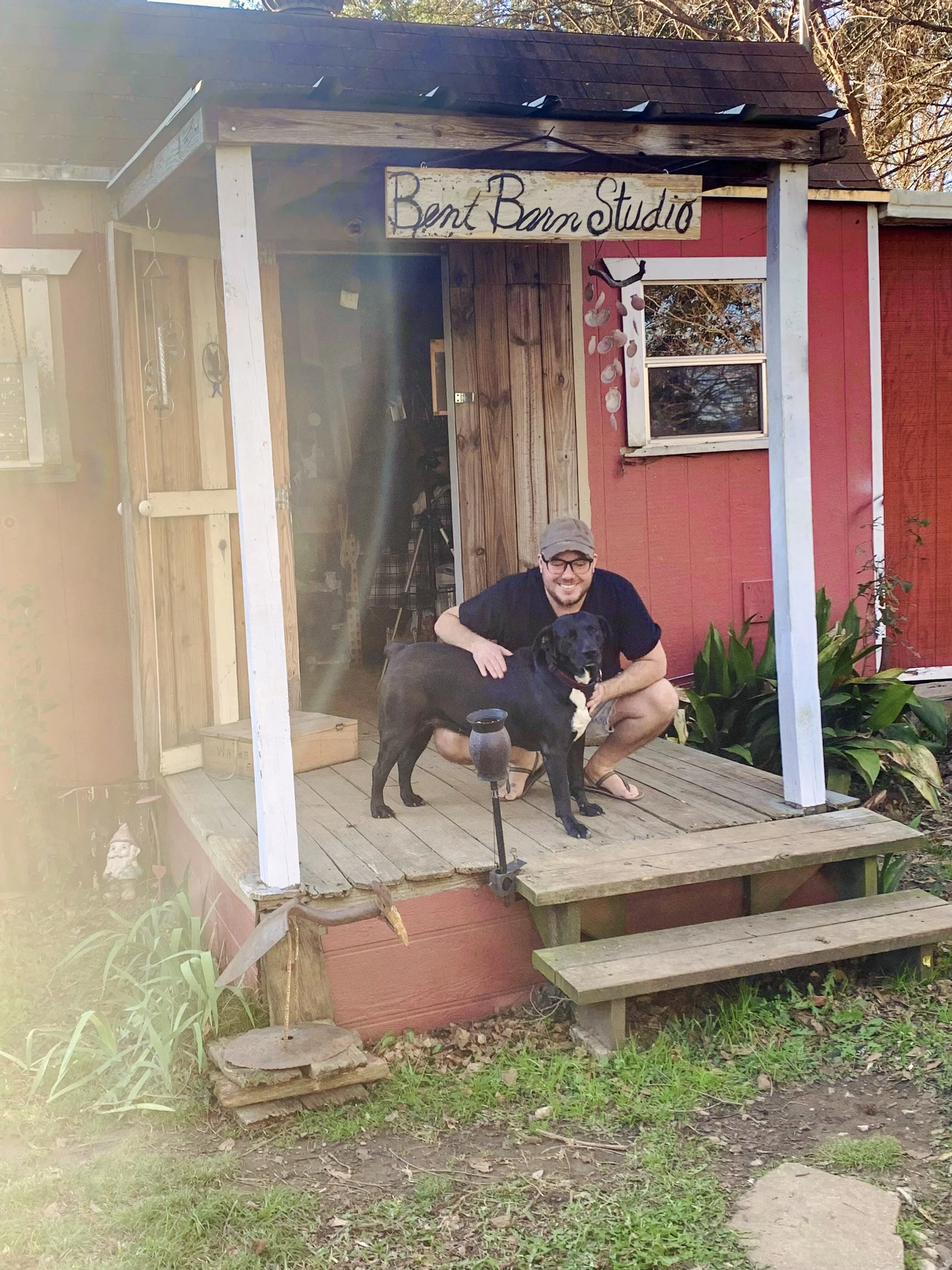A man wearing a tan cap and glasses crouches on a small wooden porch, holding a black Labrador Retriever. The porch belongs to a small, red-painted studio with a wooden door and a sign reading 'Bent Barn Studio.' Surrounding the porch are green plants and garden decorations, with steps leading down to a grassy area.
