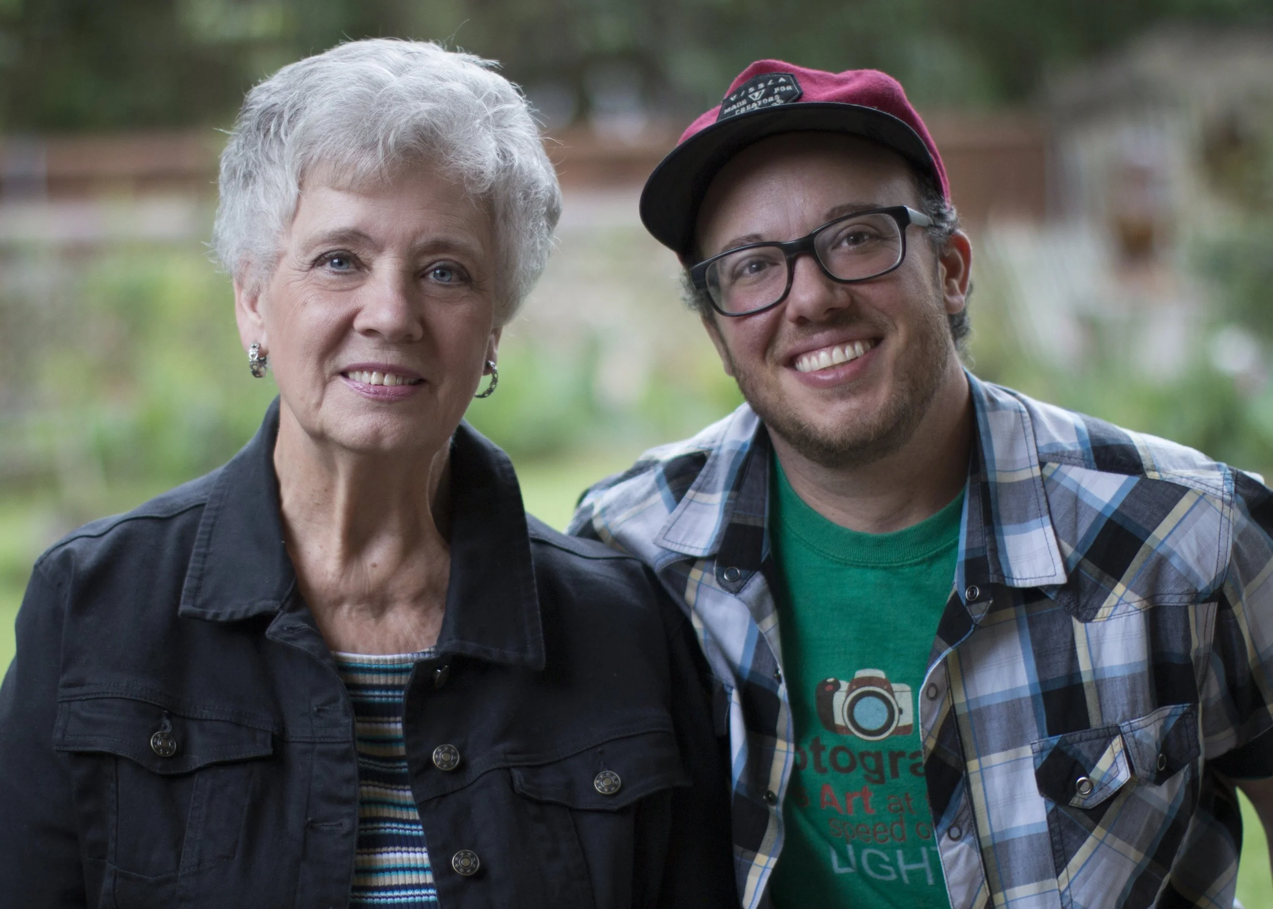 A woman with short gray hair wearing a black jacket and earrings and a young man with glasses, curly hair, a red and black cap, and a plaid shirt over a green t-shirt smiling outdoors.