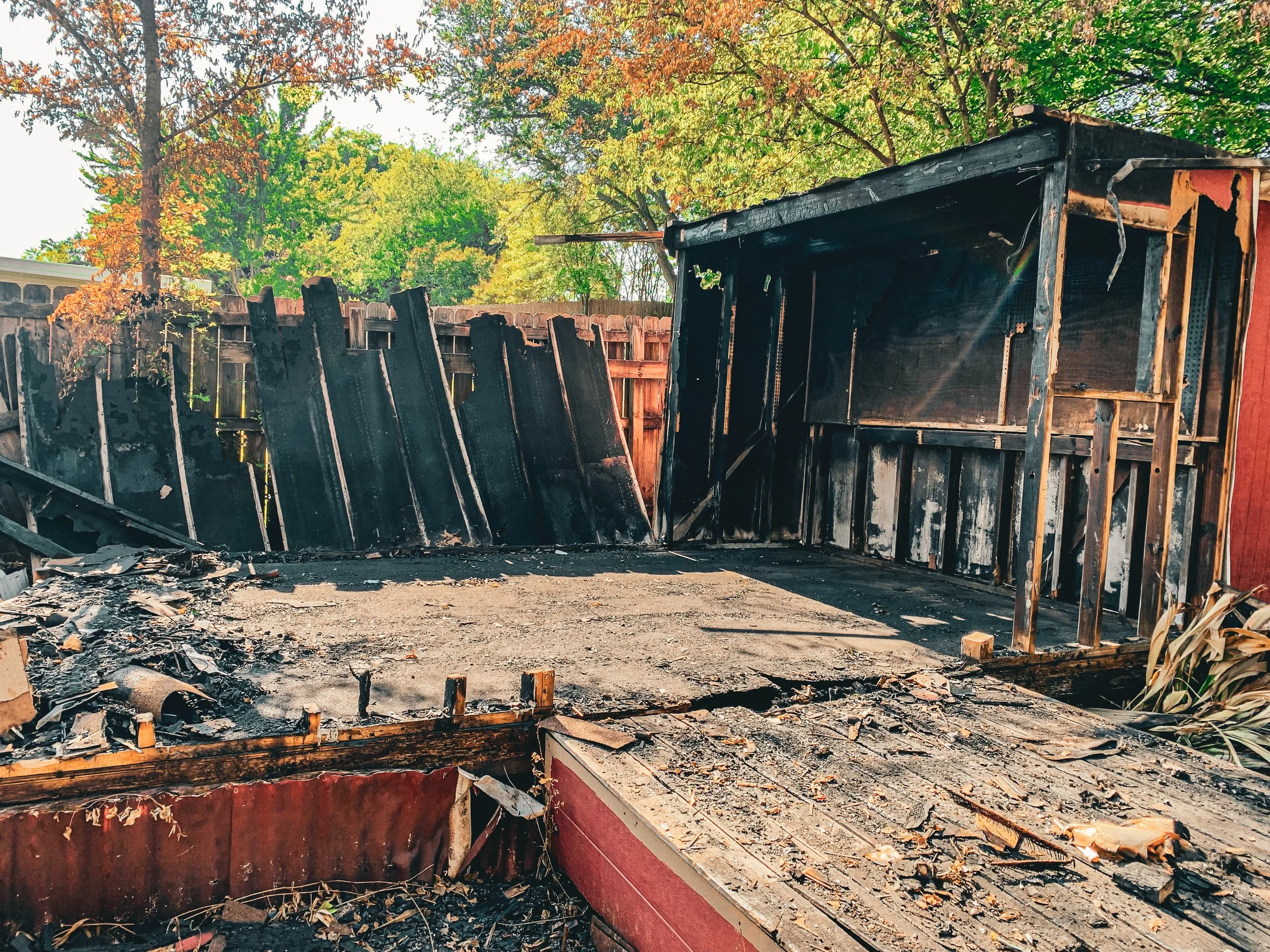 Charred remains of a burnt wooden structure in a backyard, with trees and a fence in the background.