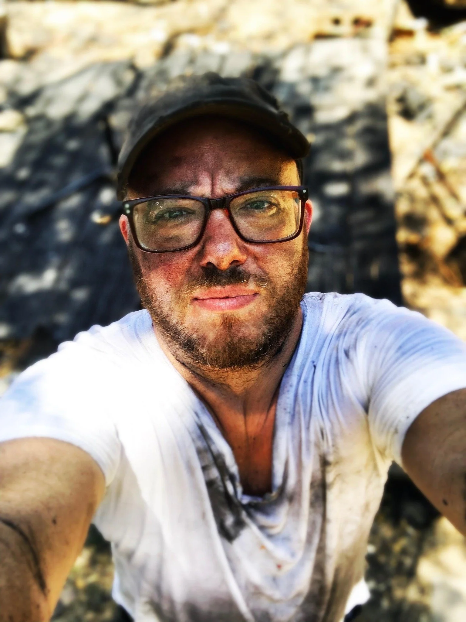 A soot-covered man wearing glasses and a black cap, with somber, tired expression and taking a selfie outdoors with rock, charred ground in the background.