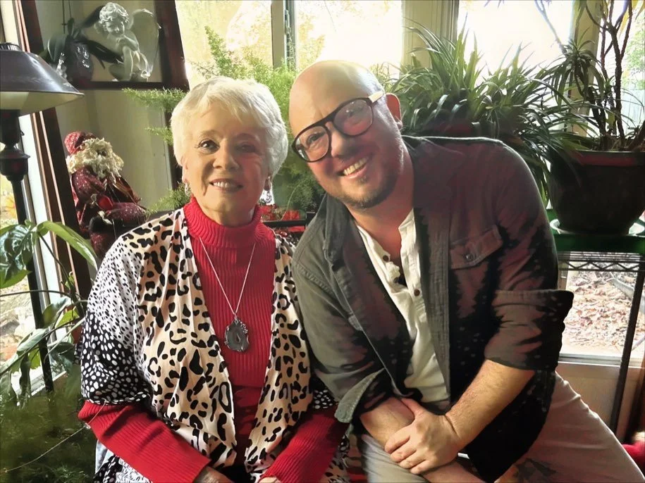 An elderly woman and a young man sitting indoors surrounded by plants, smiling at the camera.