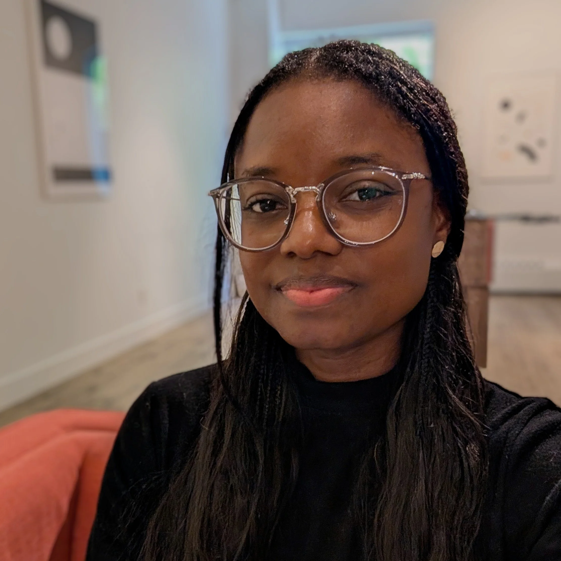 A close-up of Deneille's face with smile, wearing glasses, and with braided hair, in an indoor setting.