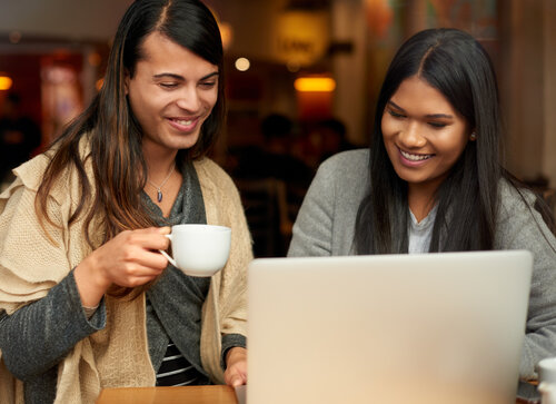 Photo Credit: Tassii/iStock/ Getty Images. Two young friends have a coffee while using a laptop together at a cafe.