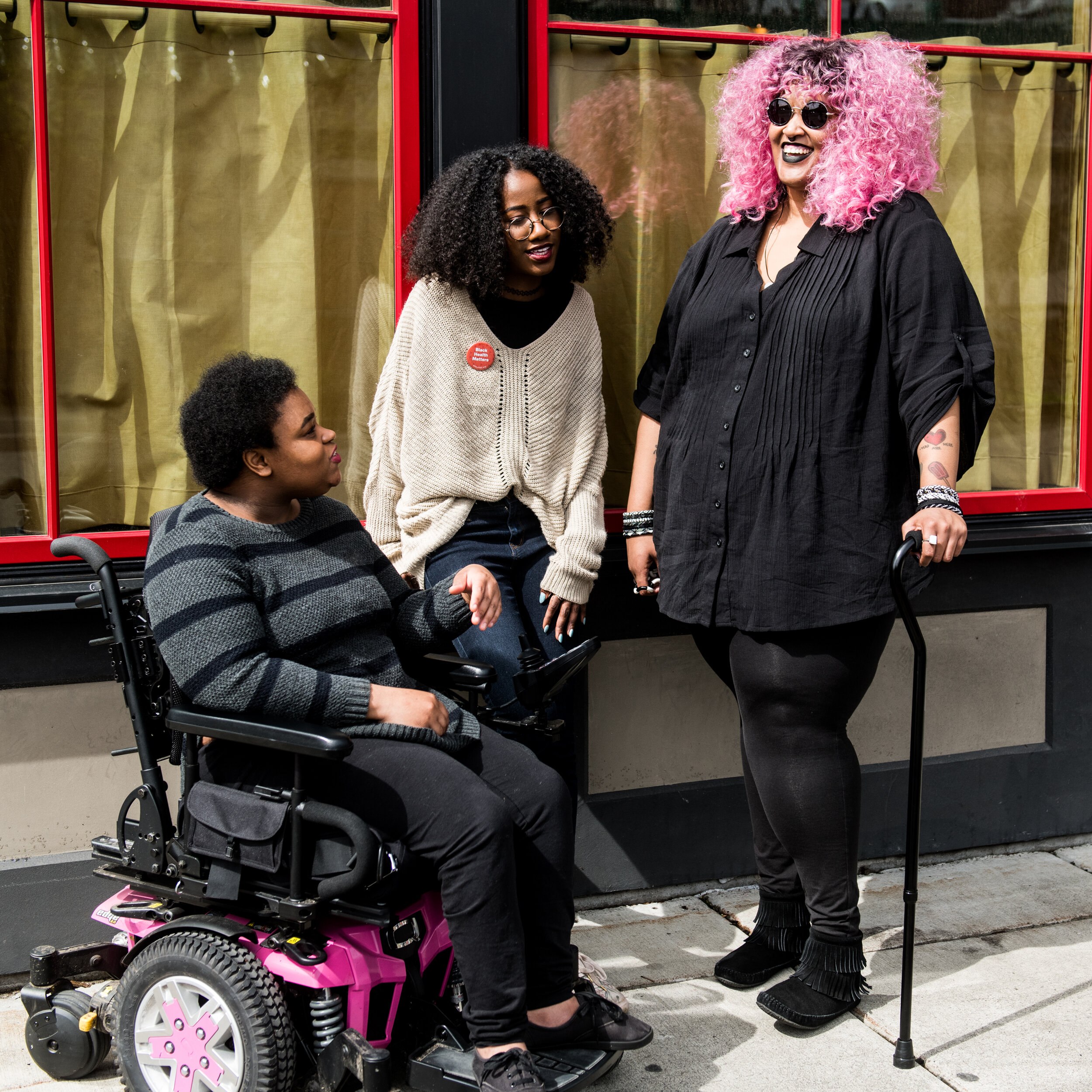 Photo Credit: Disabled And Here. https://affecttheverb.com/disabledandhere Photo taken by Chona Kasinger. “Three Black and disabled folx (one non-binary person in a power wheelchair, one woman leaning against a wall, and one non-binary person standing with a cane) engaged in conversation. All three are outdoors and in front of a building with two large windows.” N.B. Some people describe themselves using identity-first language and others use person-first language. This caption is a quotation from the photo source.