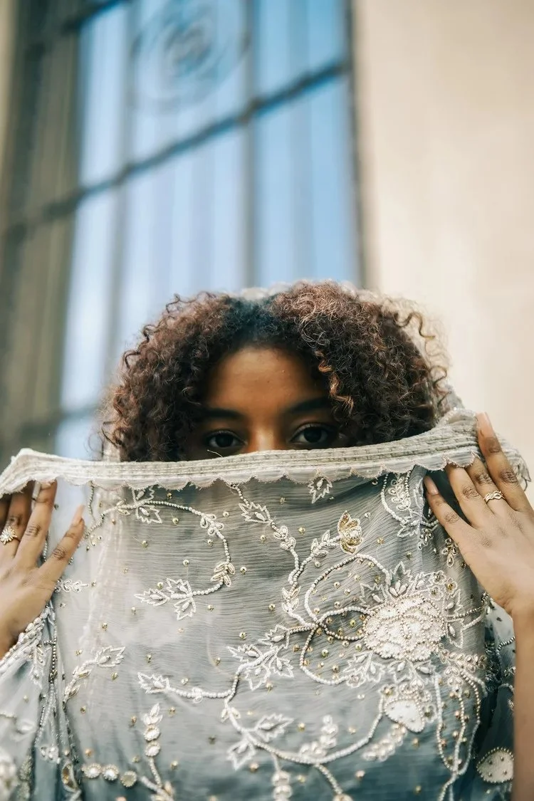 A woman holds delicate embroidered fabric across her face beside a window in A Collected Culture Pt.2. Photography by Yonathan Mesfun.