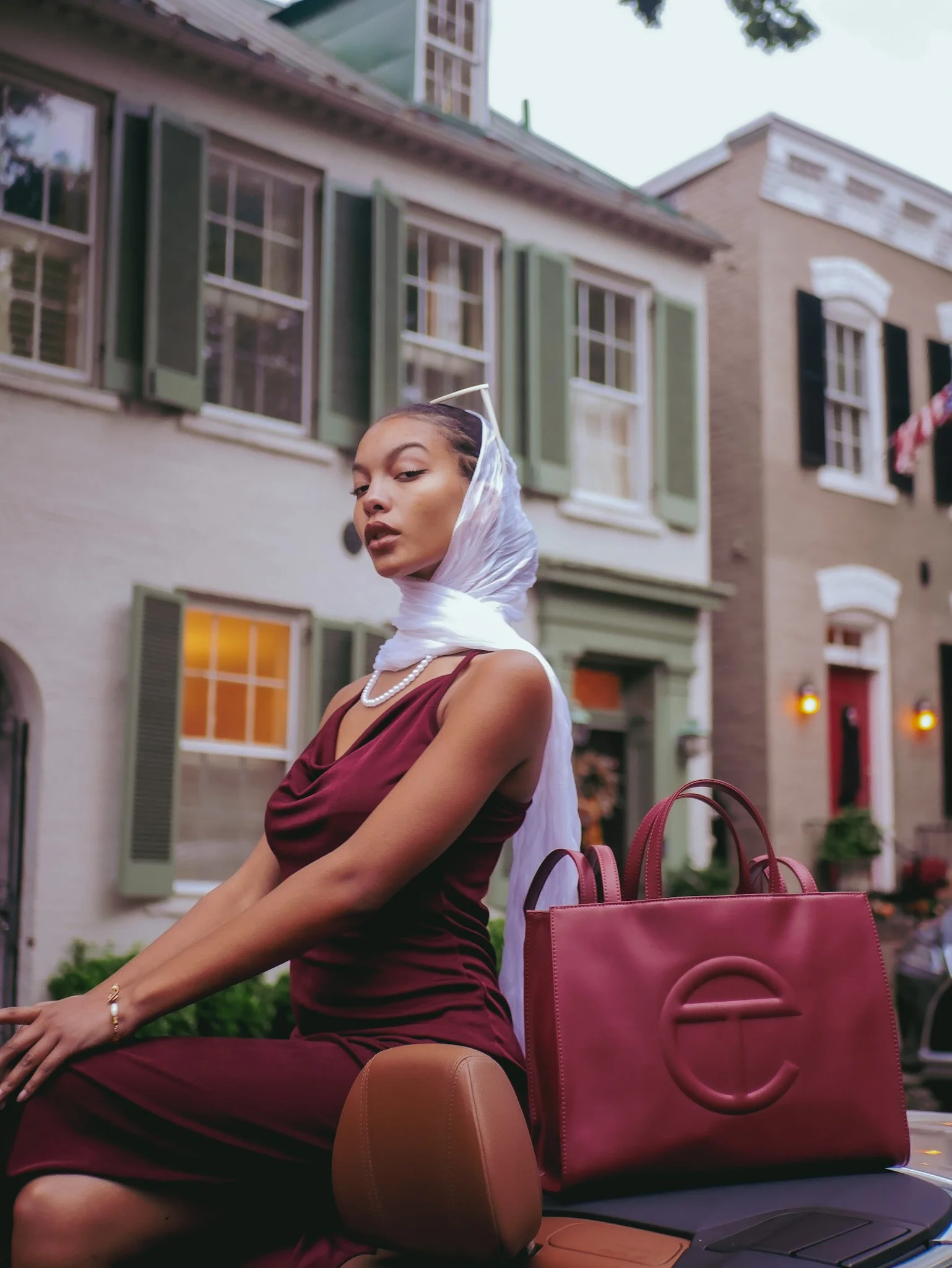 Model in a burgundy dress and headscarf beside a matching Telfar bag on a neighborhood street. Photography by Yonathan Mesfun.