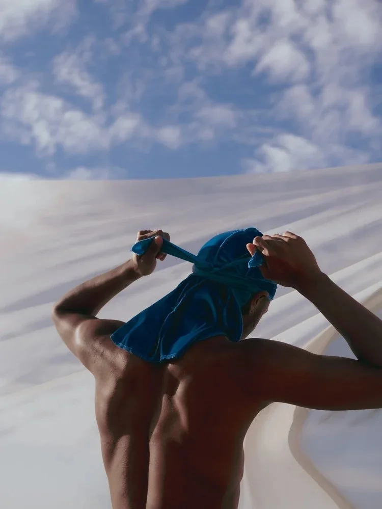 A figure ties a blue durag in front of white fabric beneath a cloudy sky in Durag Drag. Photography by Yonathan Mesfun.