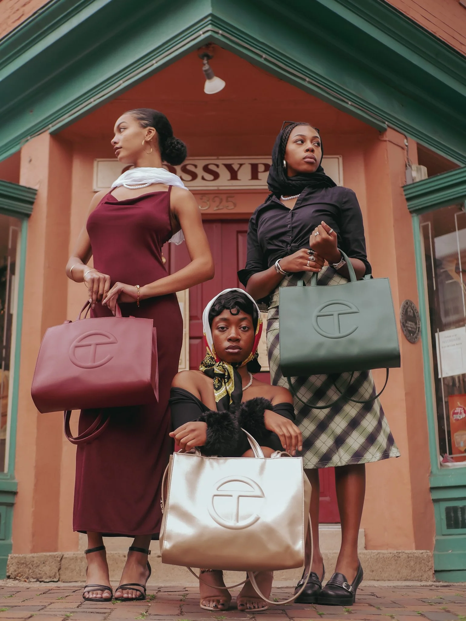 Three models at a colorful storefront holding burgundy, green and cream Telfar bags. Photography by Yonathan Mesfun.