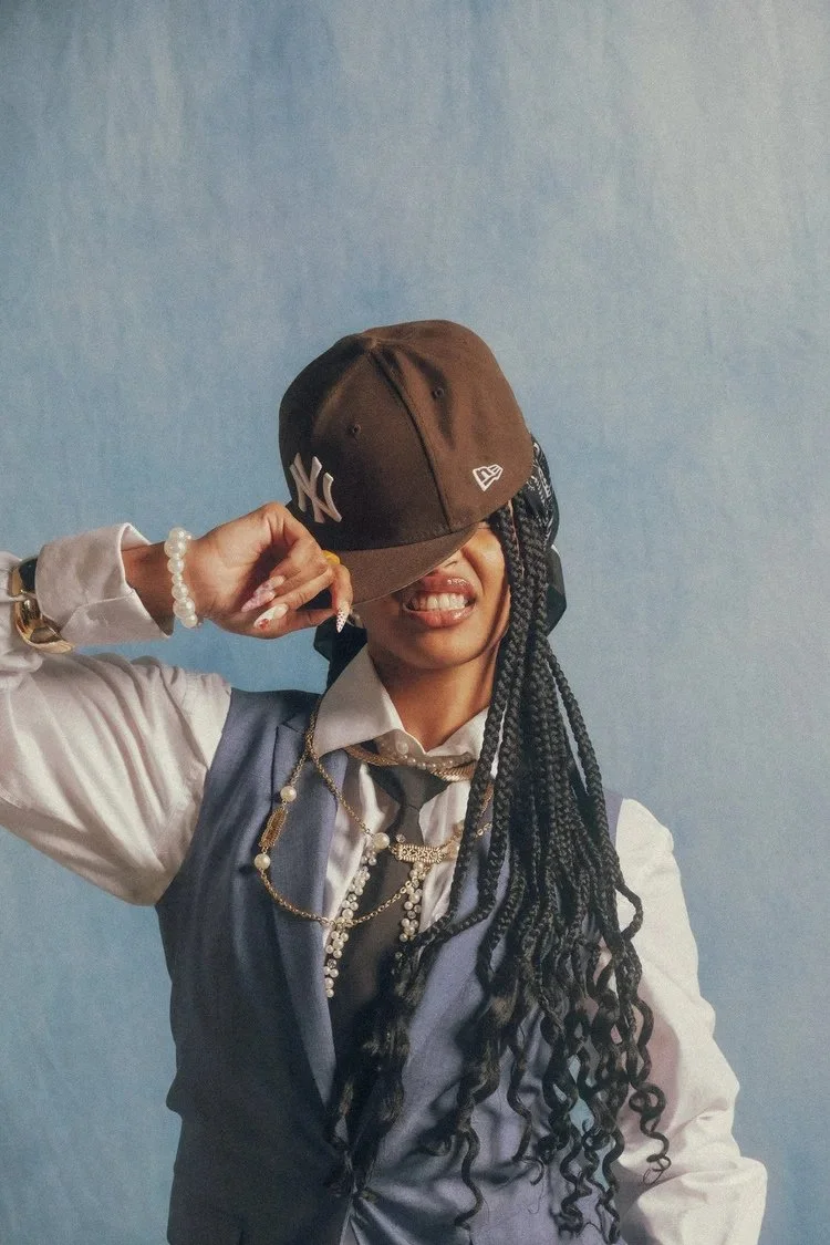 Model with long braids tips a brown baseball cap in Polos and Varsities. Photography by Yonathan Mesfun.