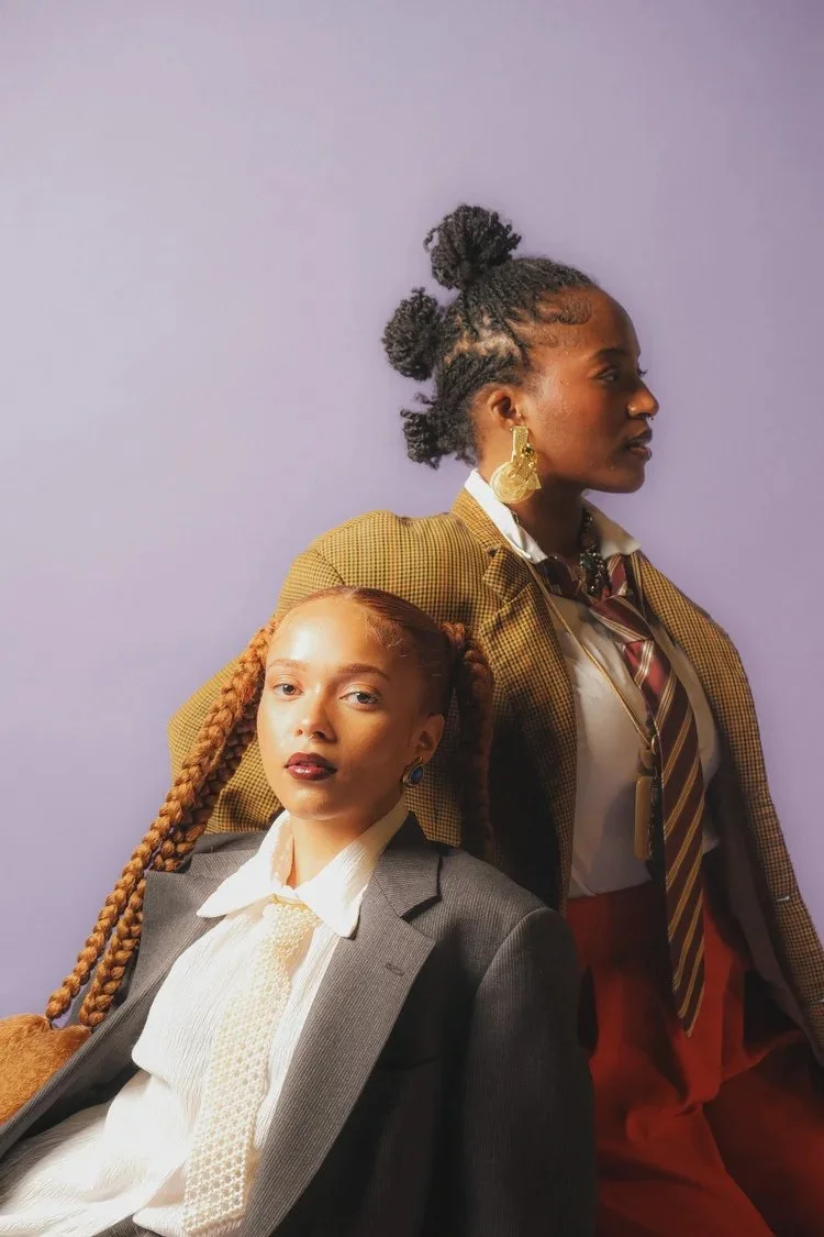 Two women with braided hairstyles wear layered shirts and oversized jackets in Sista Sista. Photography by Yonathan Mesfun.