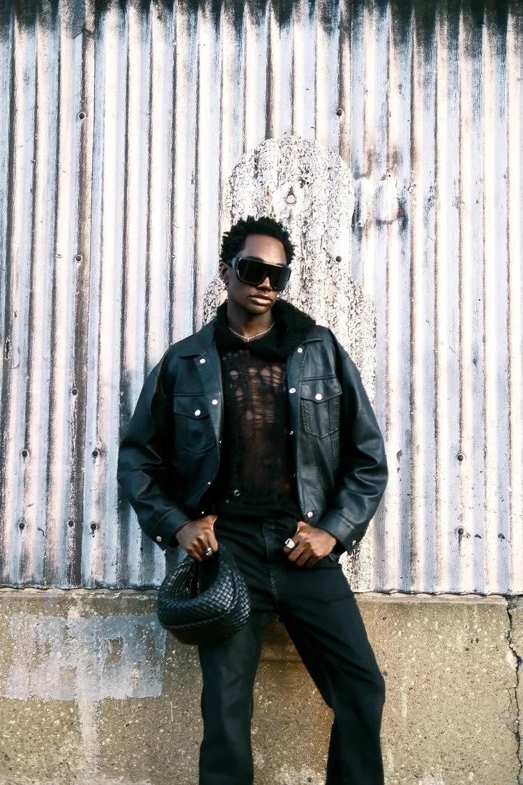 Ricky Thompson poses in black denim with a woven bag against corrugated metal. Photography by Yonathan Mesfun.