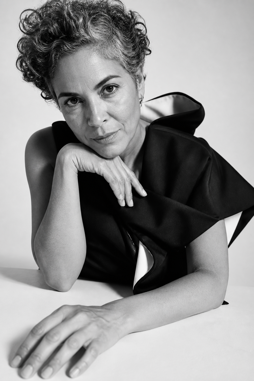 Black and white portrait of a woman with short curly hair, resting her chin on her hand, wearing a black outfit with a large collar, looking directly at the camera.