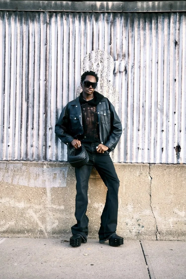 Ricky Thompson in black denim and sunglasses stands beside corrugated metal. Photography by Yonathan Mesfun.