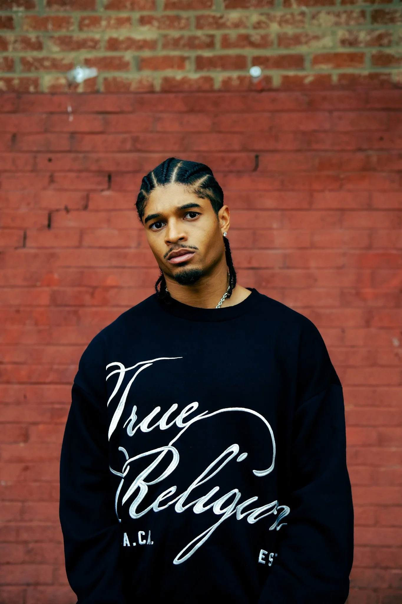 Portrait of a model in a black True Religion sweatshirt against a red brick wall. Photography by Yonathan Mesfun.