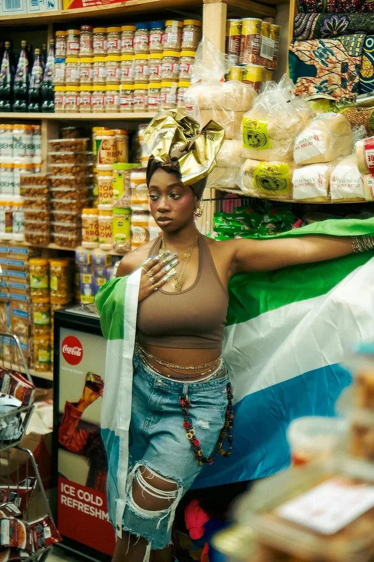 Model in a gold headwrap holds a green, white and blue flag in a grocery aisle in Sierra Leone. Photography by Yonathan Mesfun.