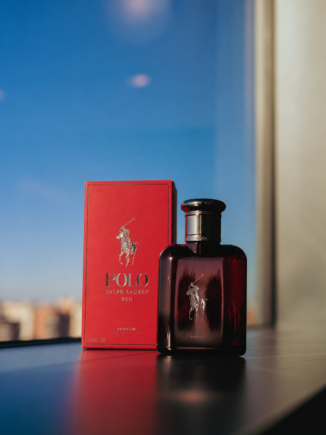 A red Ralph Lauren Polo fragrance bottle and box stand on a windowsill overlooking the city.