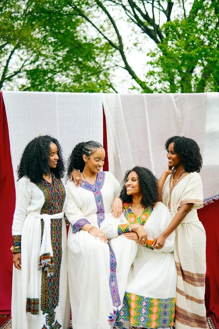 Four women in white dresses talk and smile beneath hanging textiles in Fasika. Photography by Yonathan Mesfun.