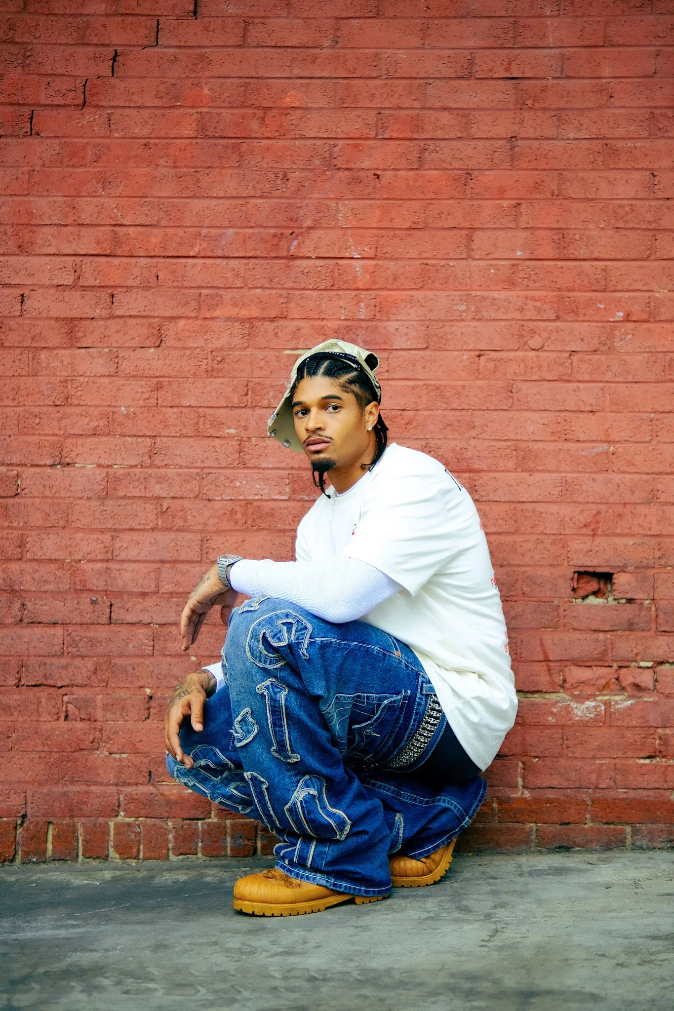 Model crouching beside a brick wall in embroidered True Religion jeans and a white shirt. Photography by Yonathan Mesfun.