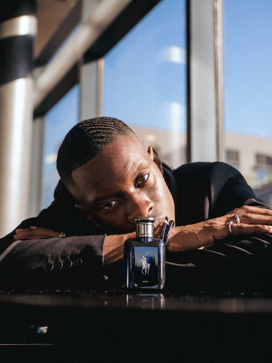 A man rests his head on folded arms beside a fragrance bottle near sunlit windows.