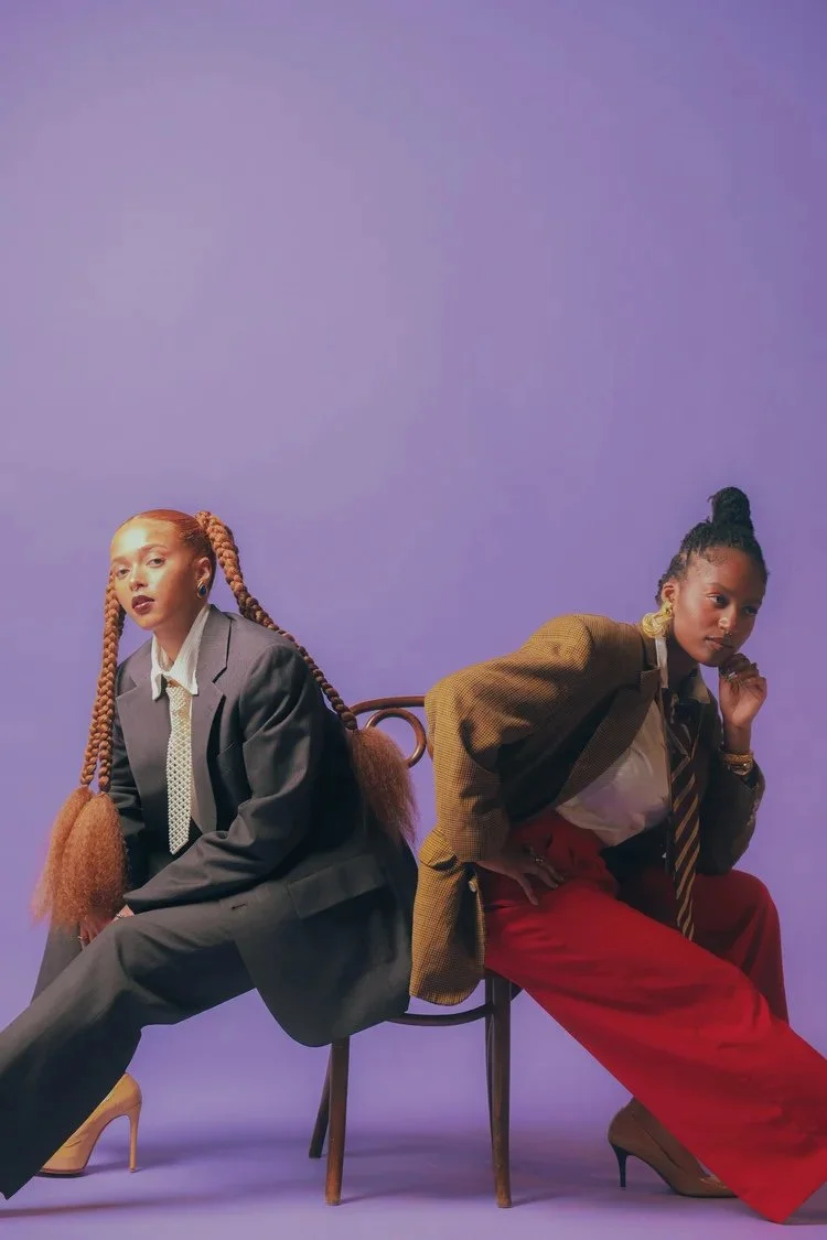 Two women share a chair in oversized suits against a purple backdrop in Sista Sista. Photography by Yonathan Mesfun.