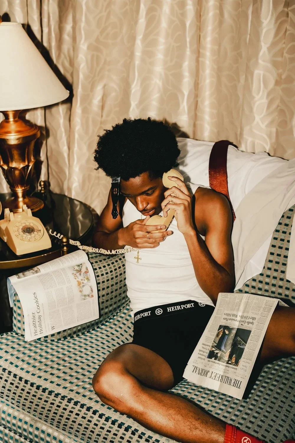 Model sitting on a bed with a corded telephone and newspaper in a Heirport campaign. Photography by Yonathan Mesfun.