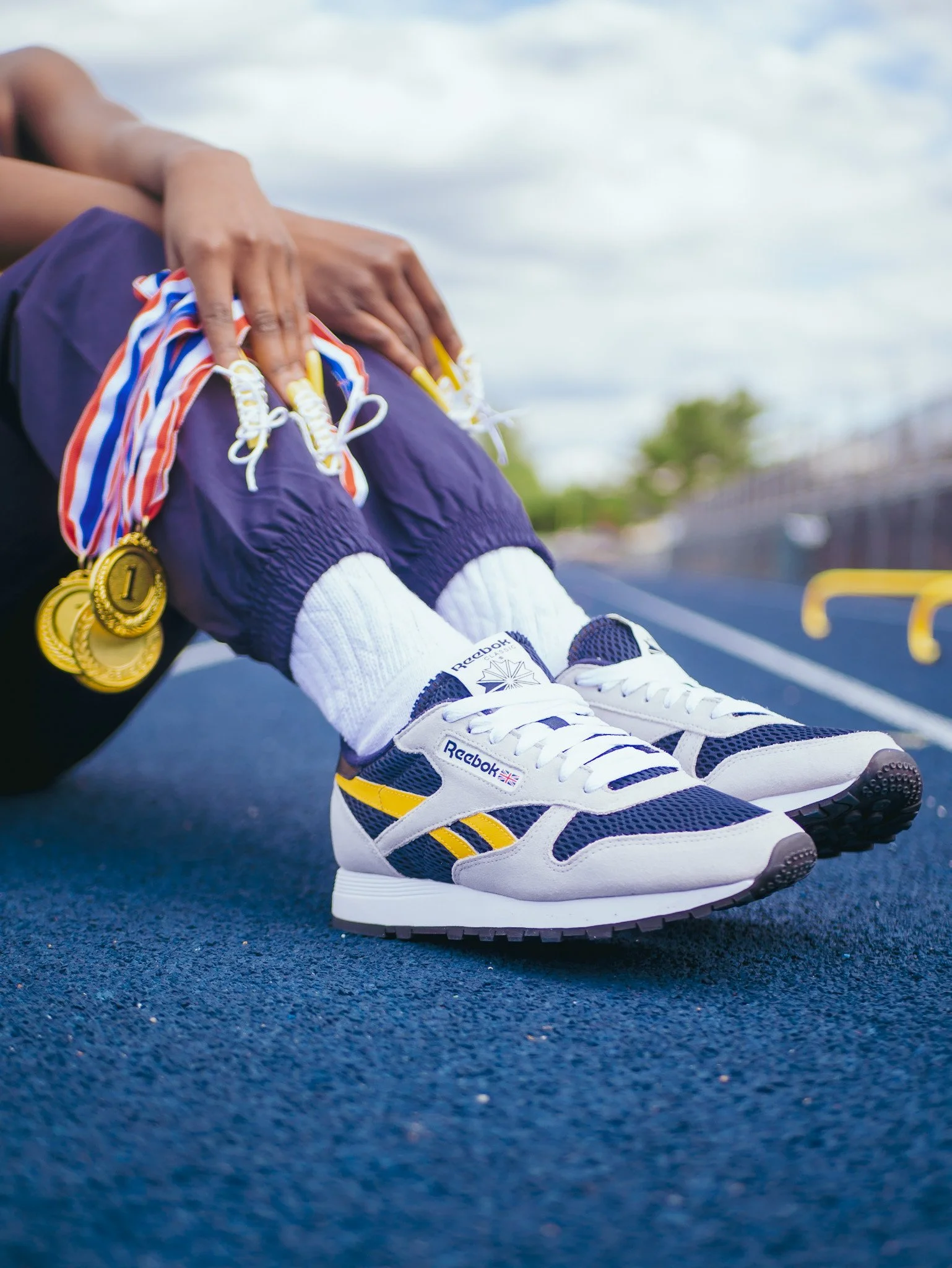 Close view of white, navy and yellow Reebok sneakers with medals on a blue sports court. Photography by Yonathan Mesfun.