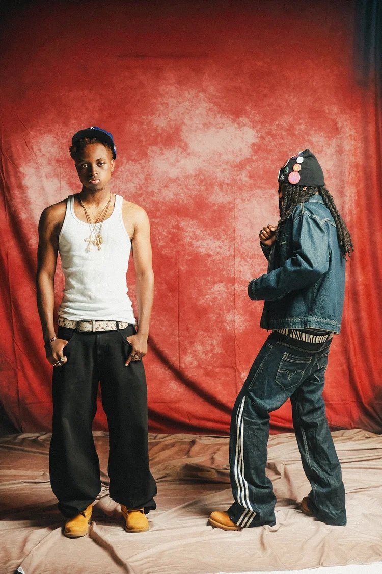 Two models in denim and a white tank top pose against a red studio backdrop in Denim Boyz. Photography by Yonathan Mesfun.