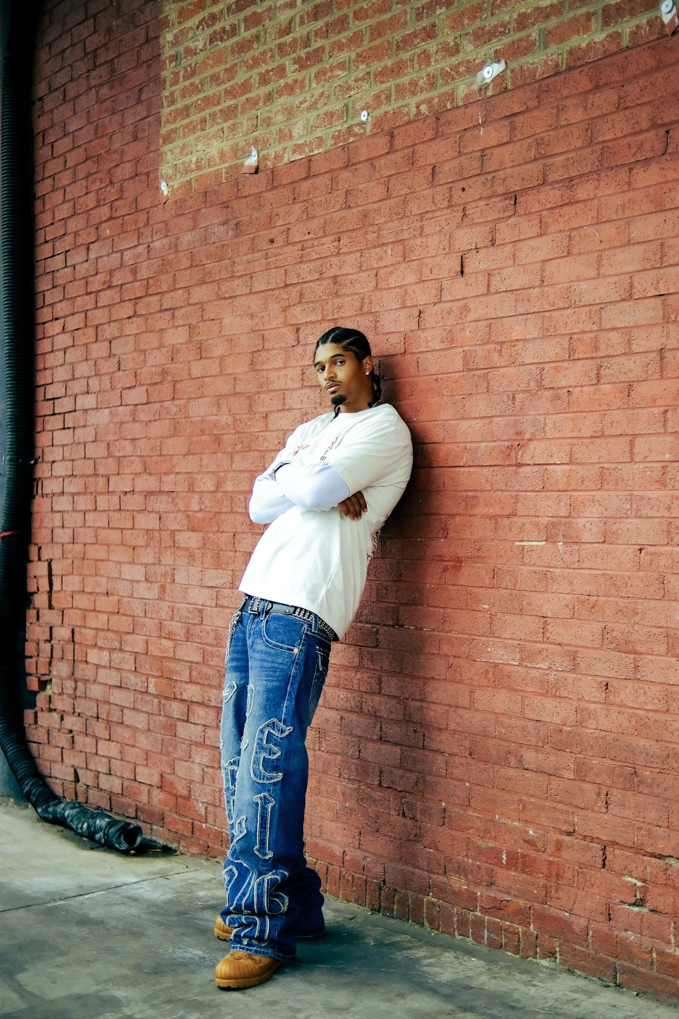 Model leaning against a red brick wall in a white shirt and True Religion denim. Photography by Yonathan Mesfun.