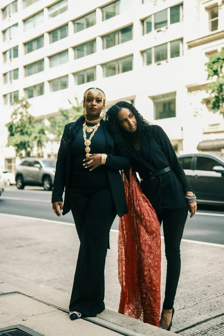 Two women in black outfits lean together on a city sidewalk in A Collected Culture Pt.2. Photography by Yonathan Mesfun.