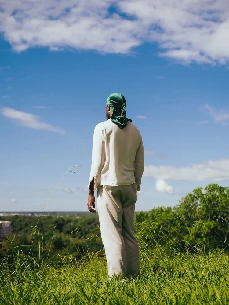 A figure in cream clothing and a green durag stands beneath a blue sky in Durag Drag. Photography by Yonathan Mesfun.