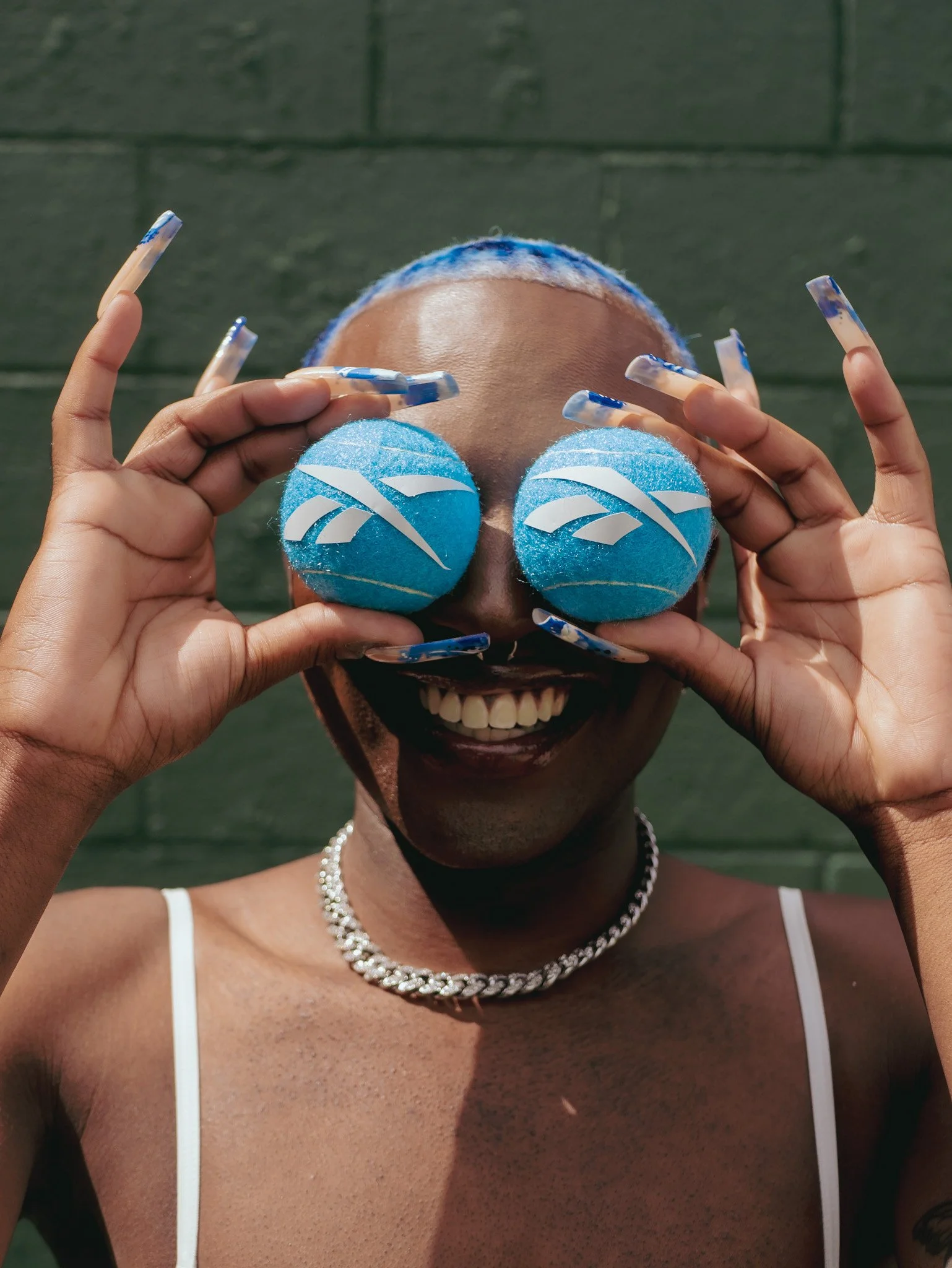 Smiling model holding two blue tennis balls over their eyes for Reebok. Photography by Yonathan Mesfun.