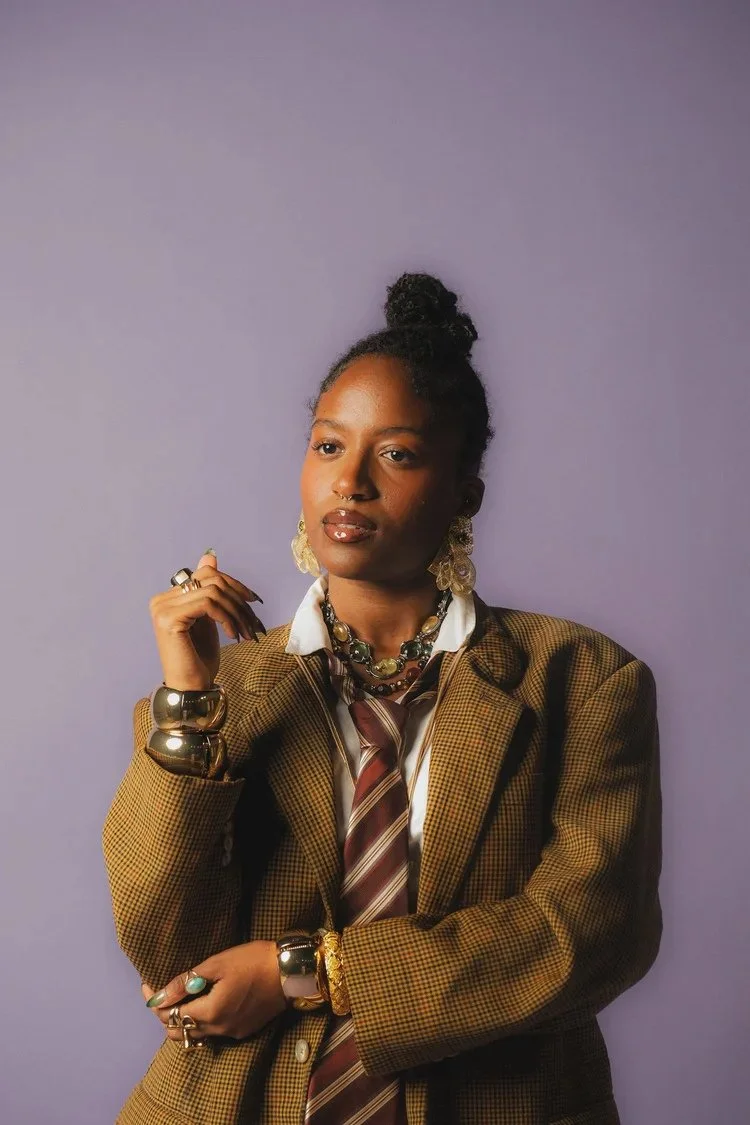 Woman in a checked jacket and striped tie poses against a purple backdrop in Sista Sista. Photography by Yonathan Mesfun.