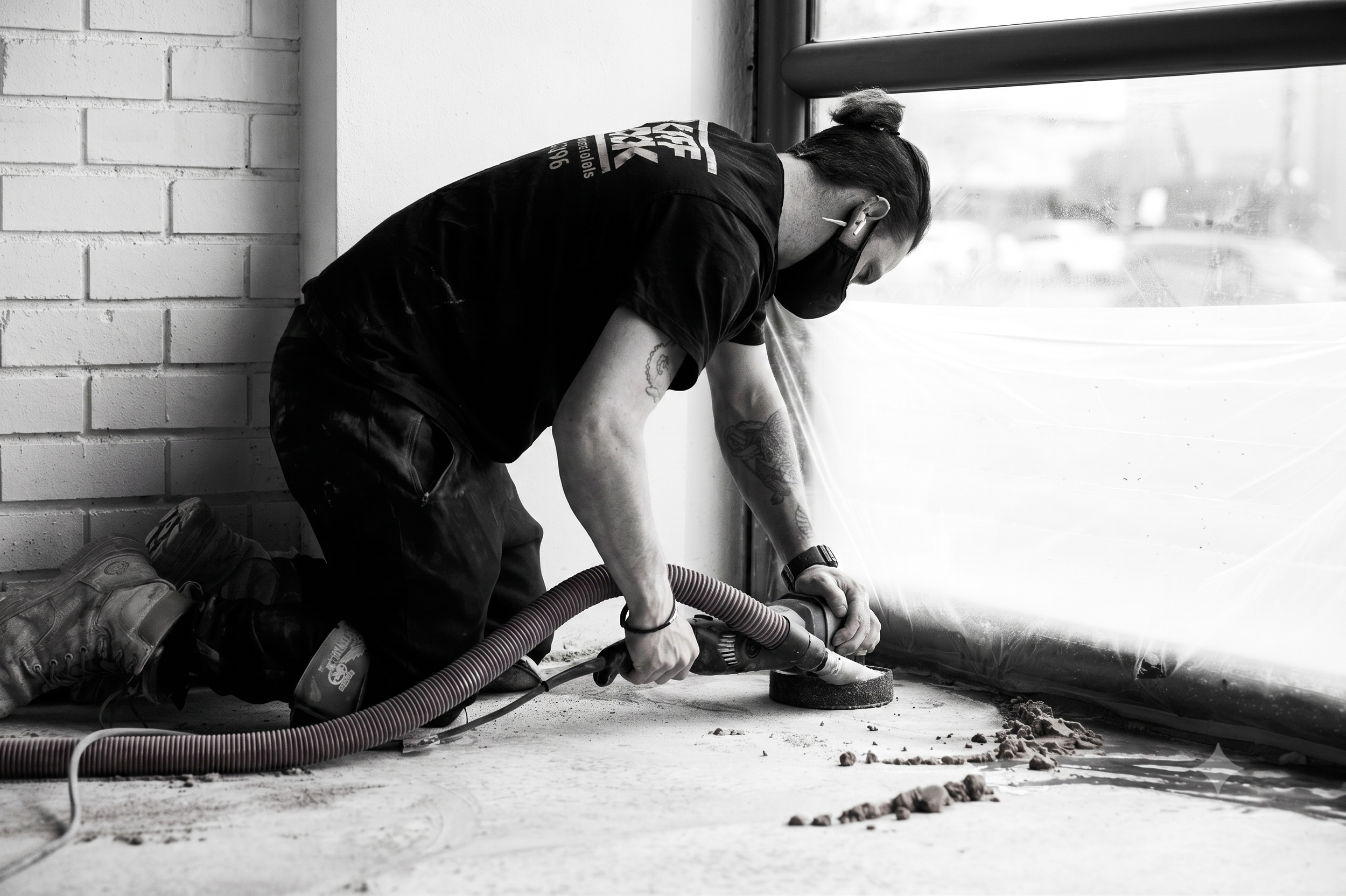 A person wearing work boots, a black mask, and protective gear is bending over, operating a large hose or pipe on a construction site by a window that shows a blurred outdoor scene.