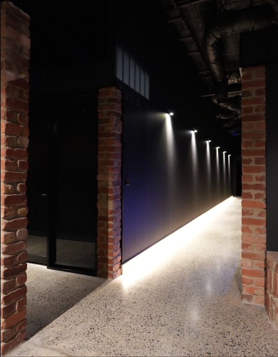 A modern hallway with exposed brick pillars, black corrugated wall, and subtle ceiling lighting, with polished concrete floors.