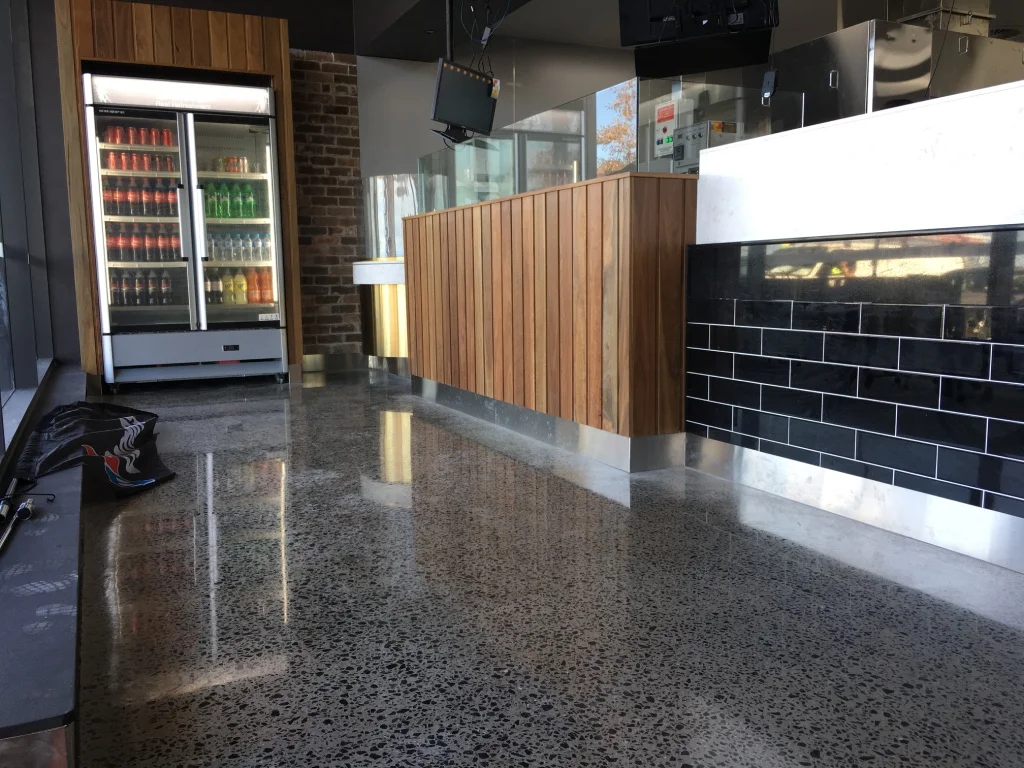 Empty fast food restaurant counter with a refrigerator stocked with drinks in the background, a cash register area with wood paneling, and black tiled walls.