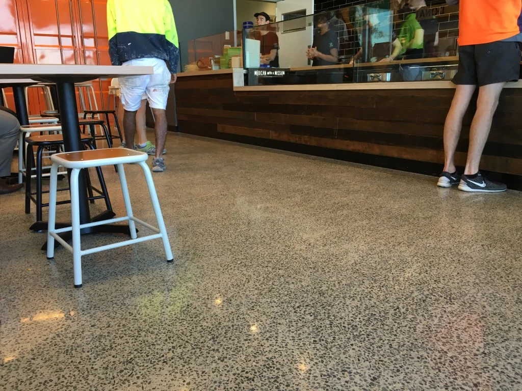 Inside a fast-food restaurant with a counter and a few customers waiting in line. There are stools and chairs, and a wooden counter with a glass partition.