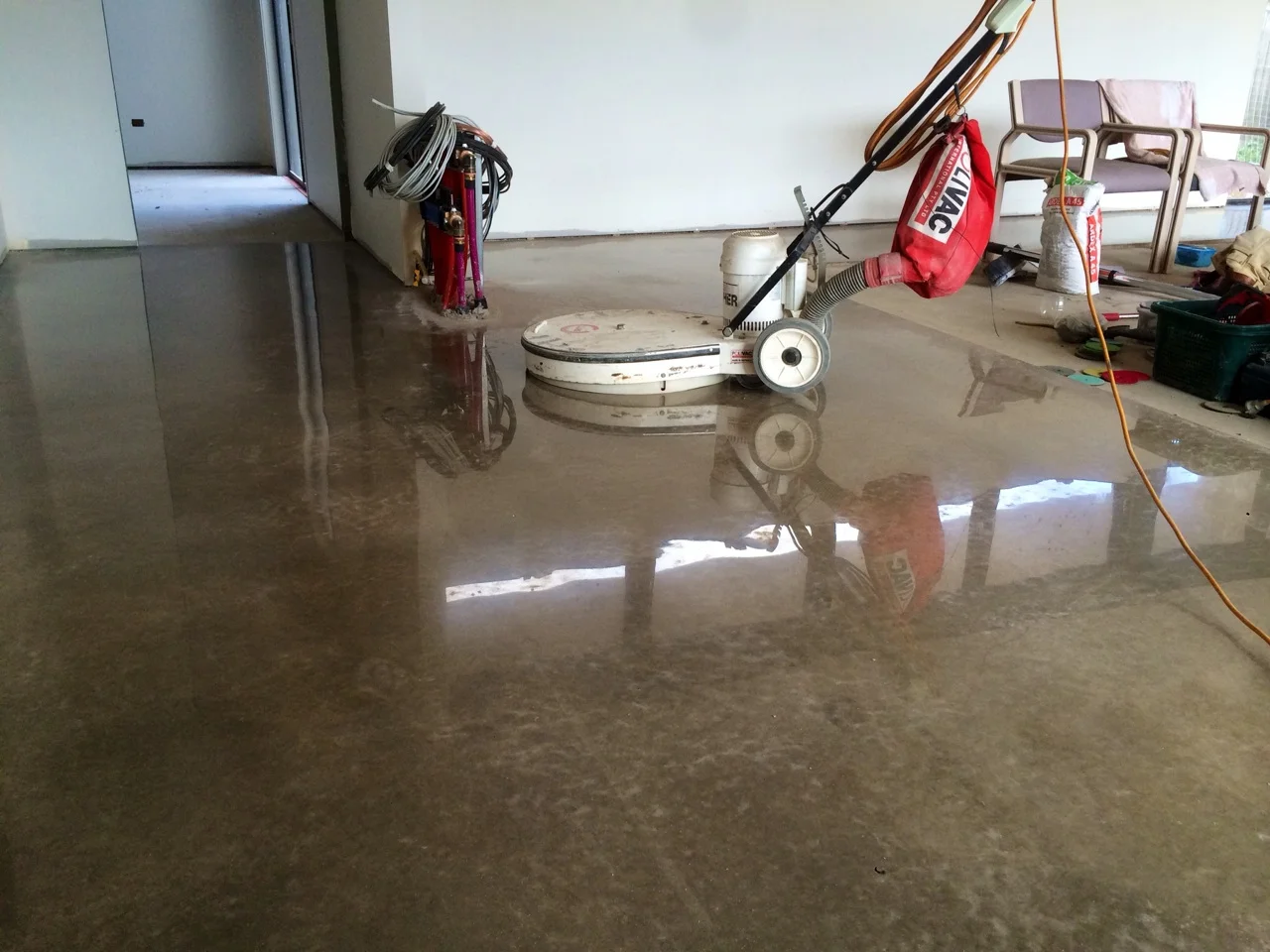 A concrete floor being polished, with a floor polishing machine, cords, and various tools and furniture scattered around a room that appears to be under renovation.