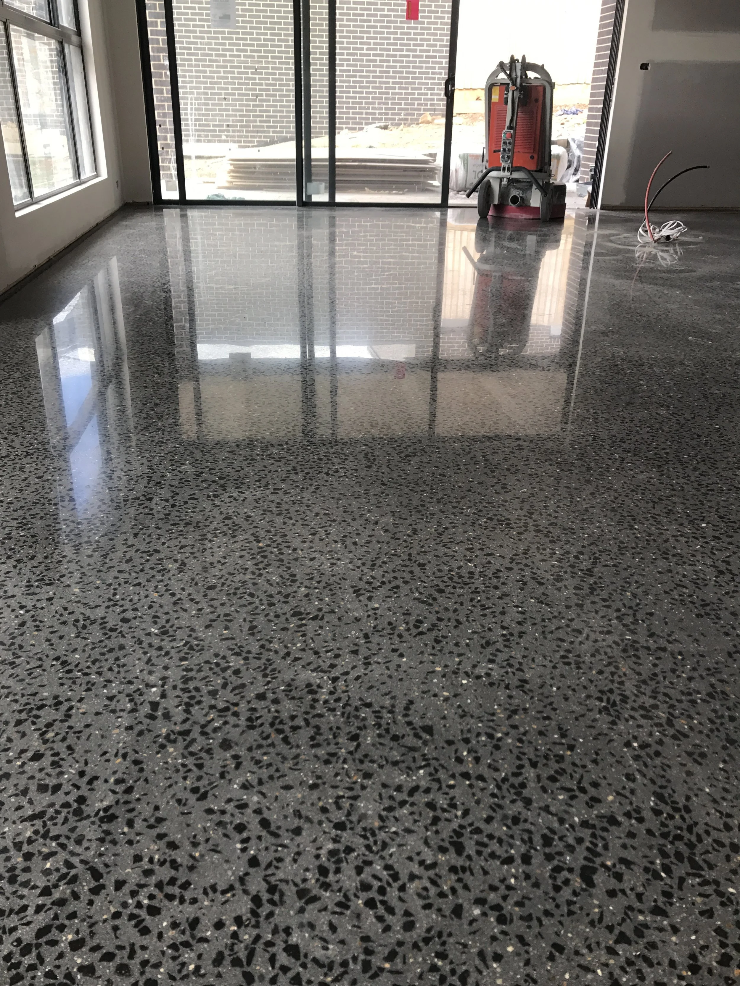 Interior space with polished terrazzo flooring, large glass sliding door leading outside, construction equipment on the floor, and an area under construction outside.