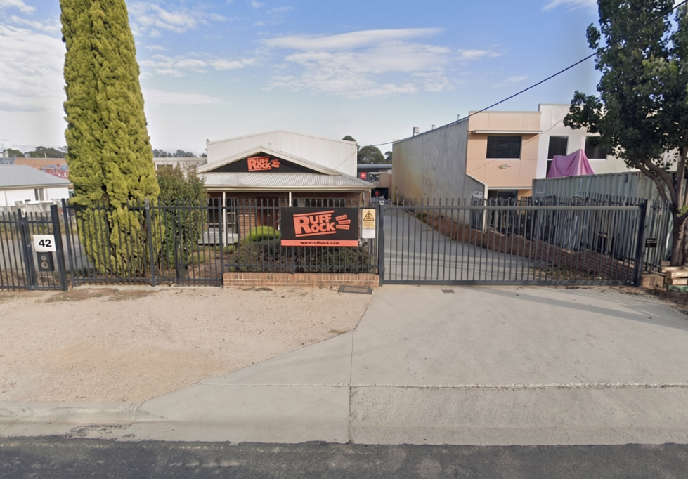Exterior view of a building with a sign that says 'Duff Rock,' a fenced driveway, and a tall tree on the left.