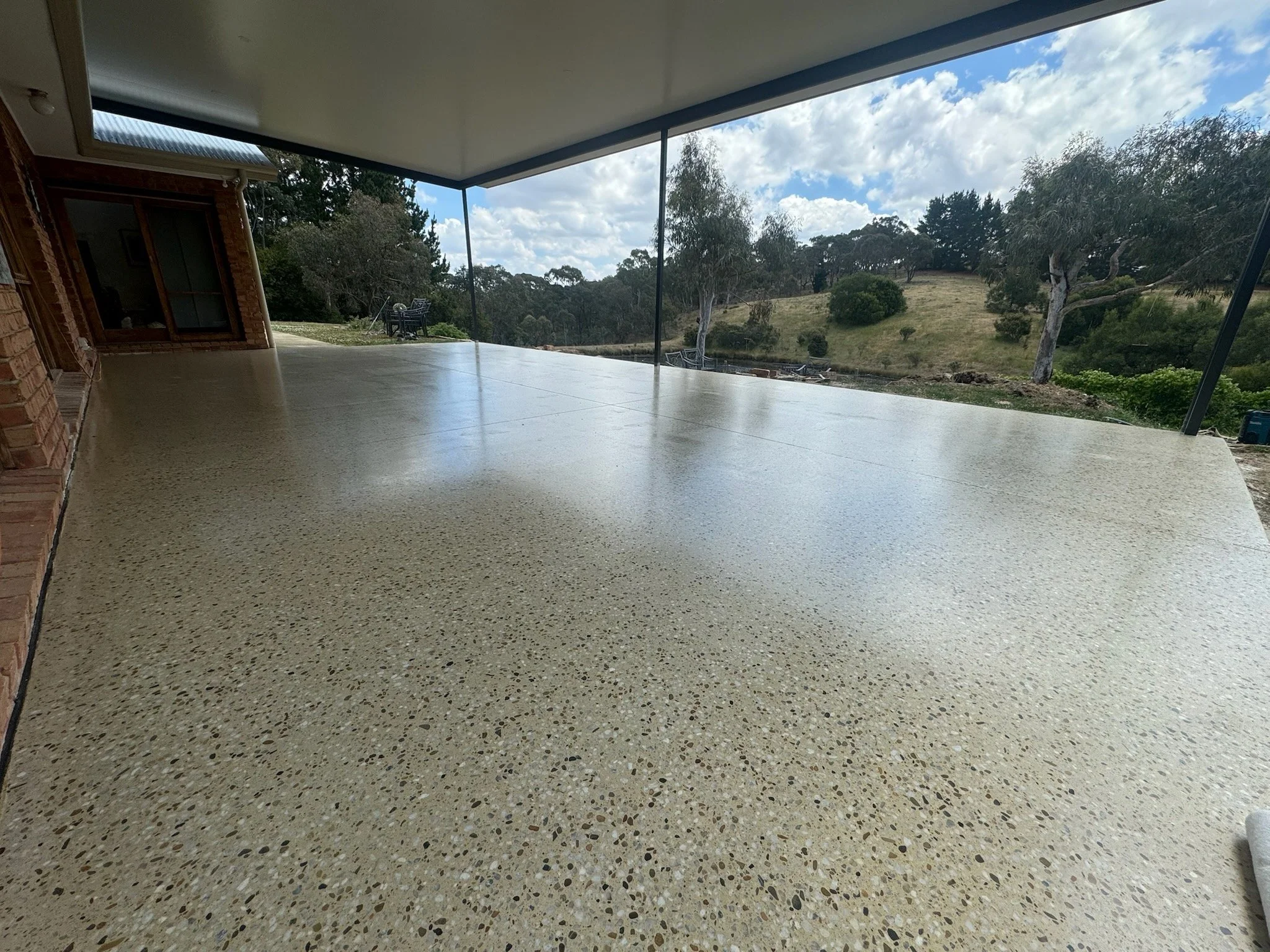 A spacious outdoor patio with a textured concrete floor, surrounded by a yard with trees and grassy hills, under a partly cloudy sky.