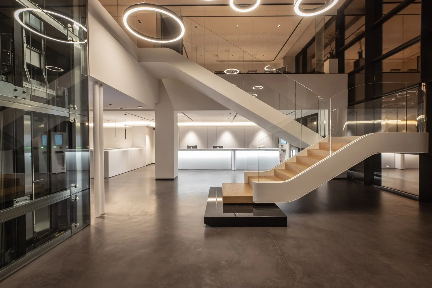 Modern hotel lobby with white reception desk, staircase with glass railing, circular ceiling lights, and black and white decor. Nuvolato Architop flooring, burnished concrete finish, high-end architectural floors. Seamless concrete overlay, micro-cem