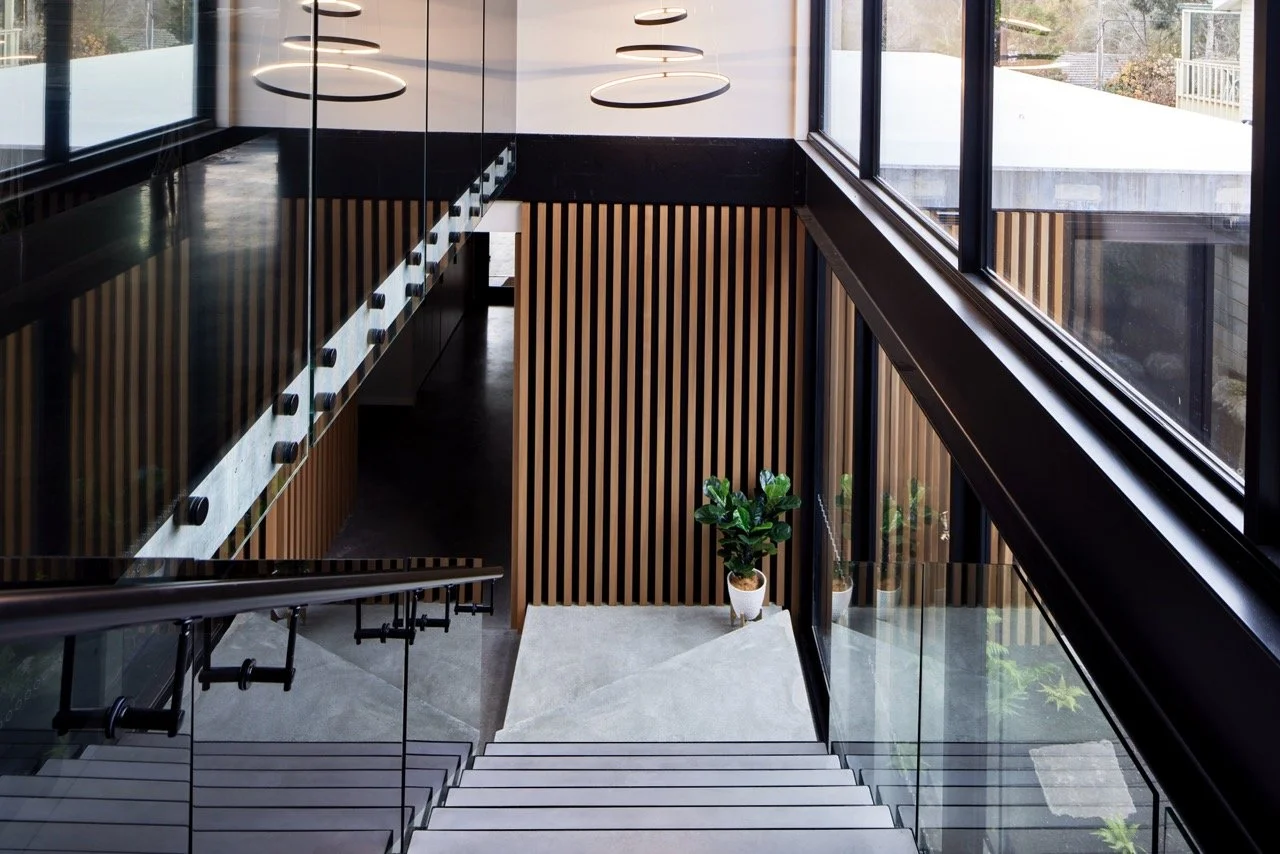 Interior view of a modern staircase in a building with large windows, wooden slat walls, and a potted plant at the bottom of the stairs.