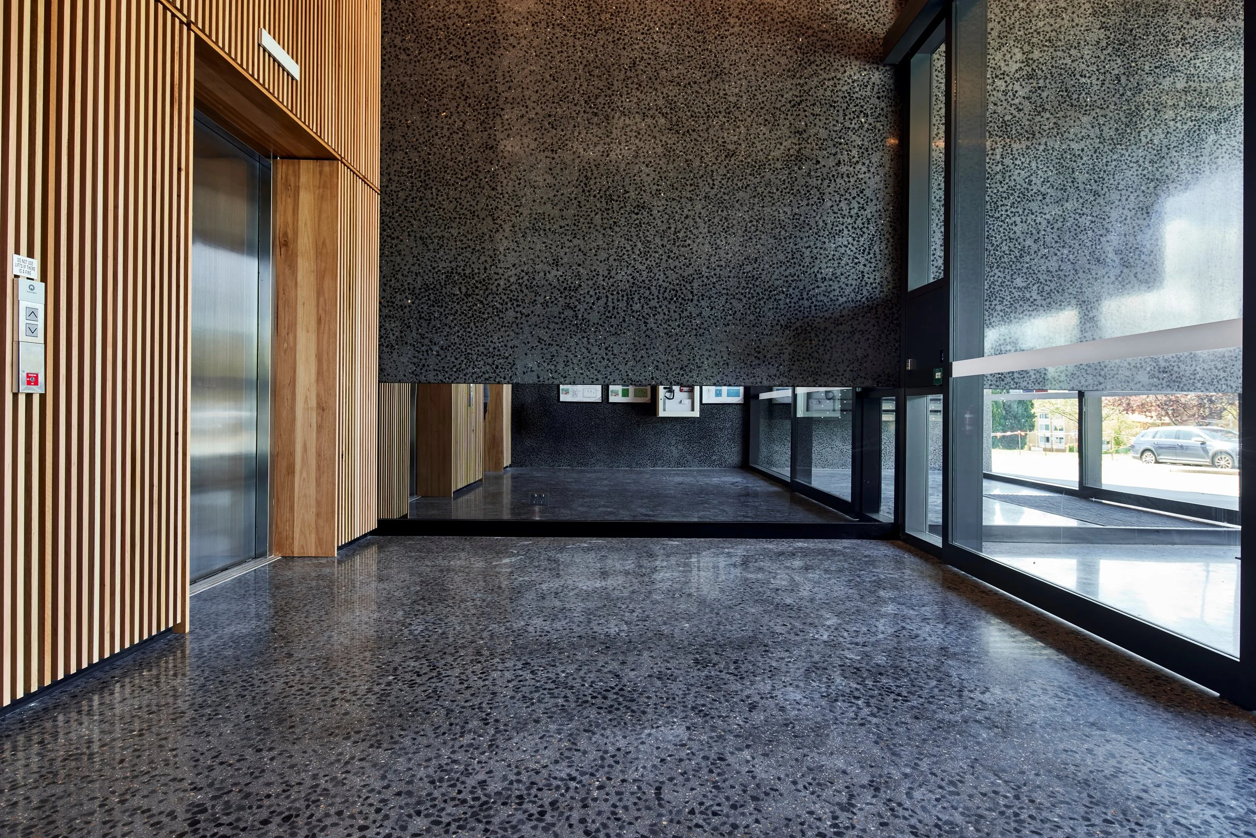 Modern building lobby with wooden slat walls, dark speckled terrazzo flooring, and large glass windows. An elevator with a panel is on the left, and the lobby has a high ceiling with natural light coming through the windows.