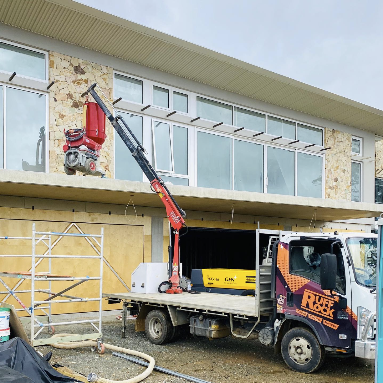 Construction site in front of a modern house with large windows, scaffolding, and a crane truck with a red jackhammer on the bed.