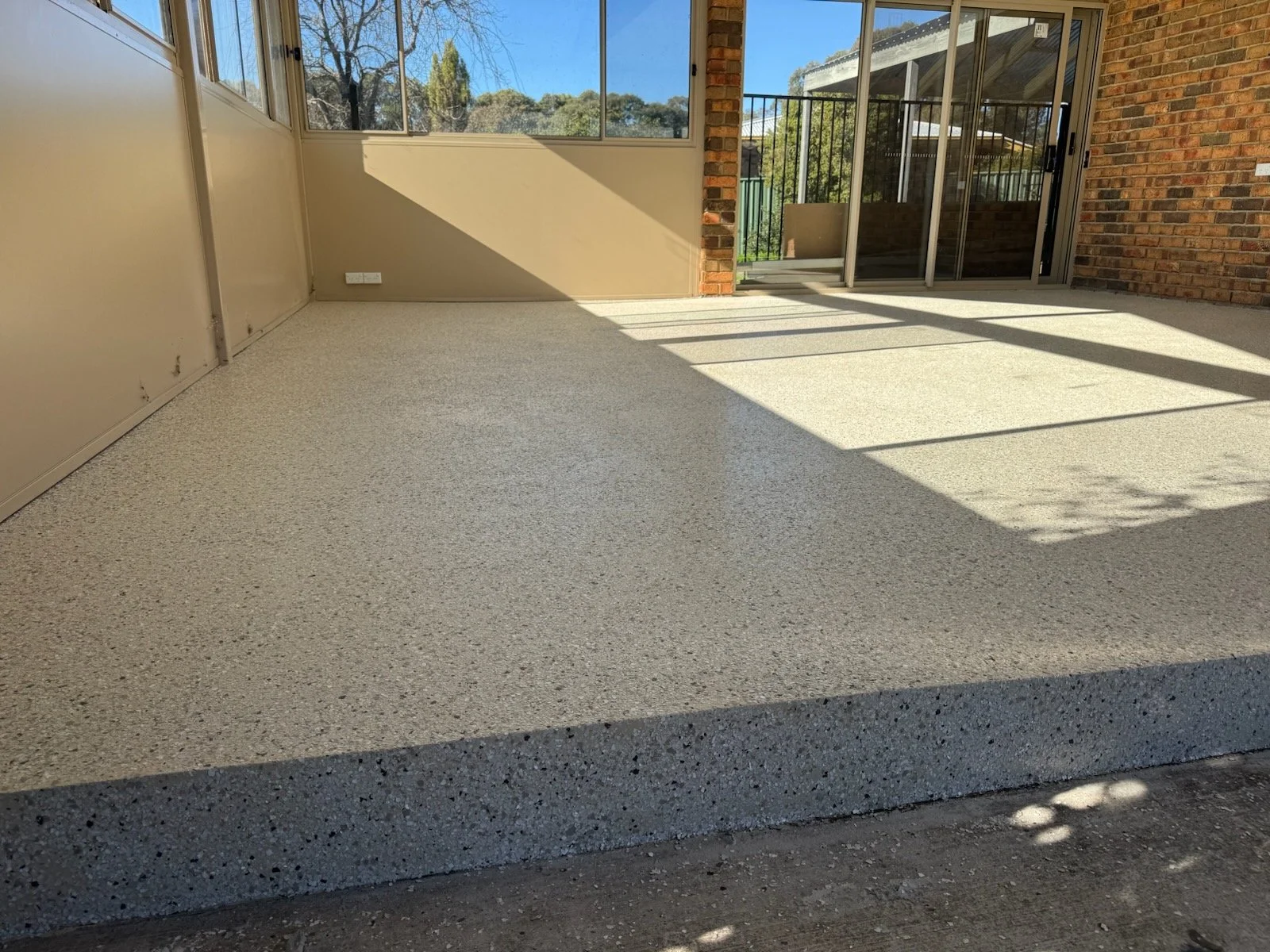 Enclosed porch with beige speckled concrete floor, large windows on left, brick wall and sliding glass door on right, and a view of trees and a patio area outside.