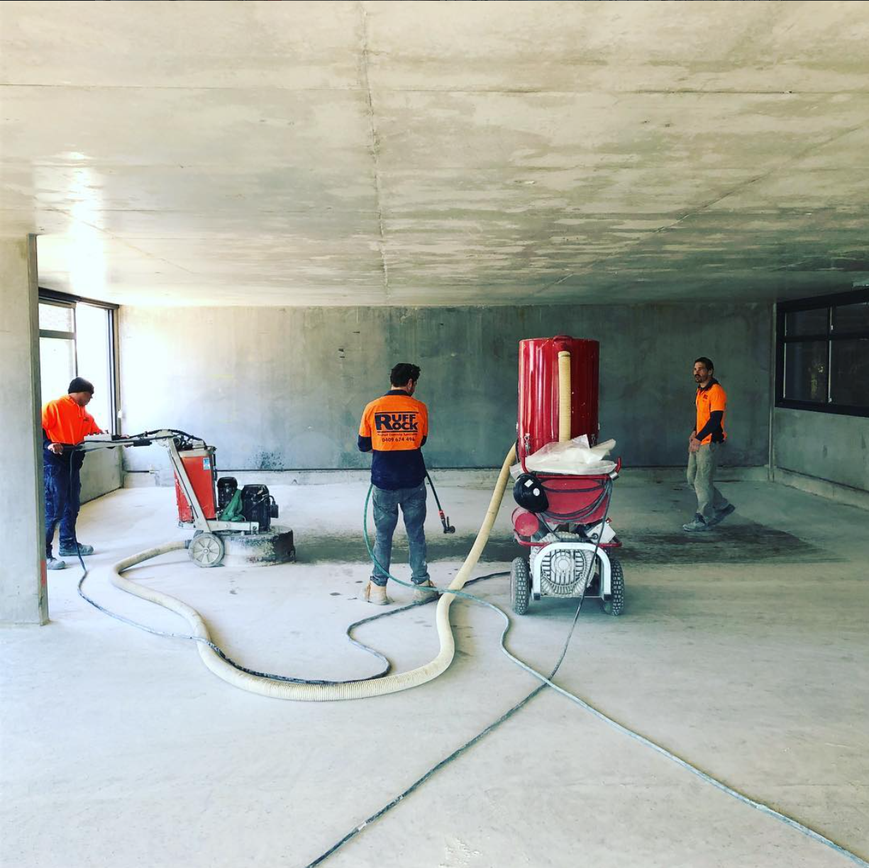 Construction workers in orange safety vests are using equipment to grind or polish a concrete surface inside a building under construction.