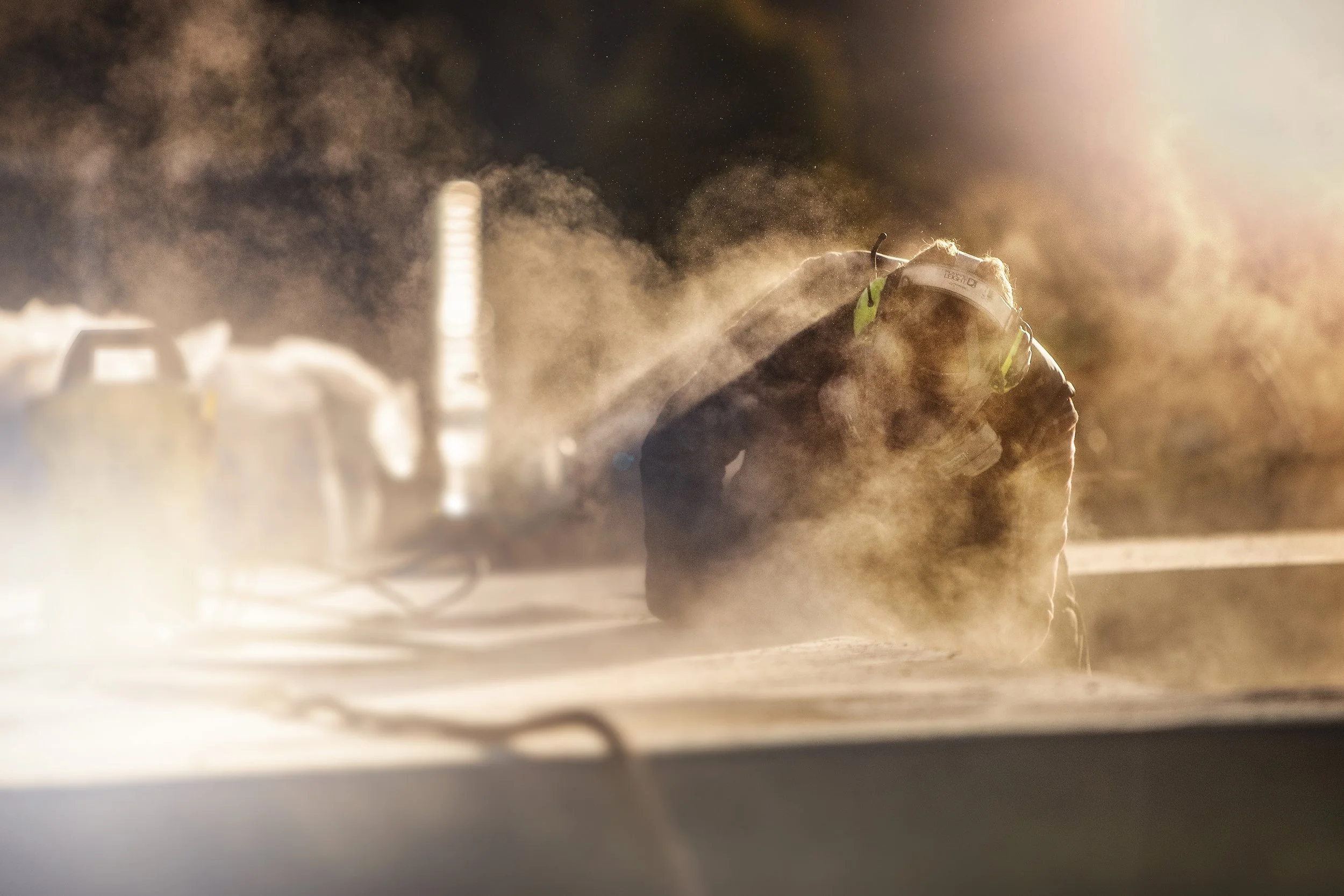 A person wearing a helmet and protective gear kneeling on a dirt surface, surrounded by dust and smoke, illuminated by bright light from the right side.