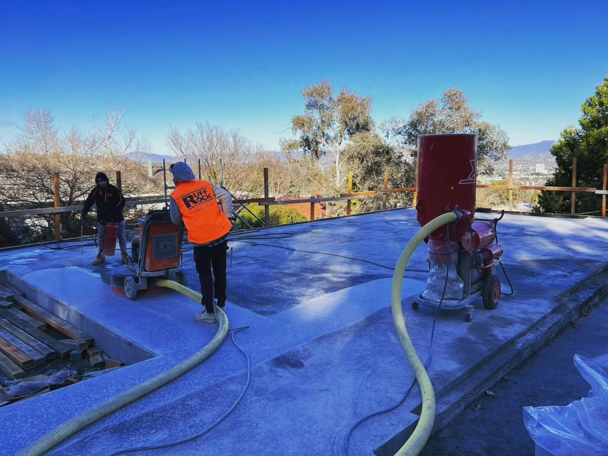 Construction workers using equipment to apply a coating or sealant to a flat roof outdoors, with trees and mountains in the background.