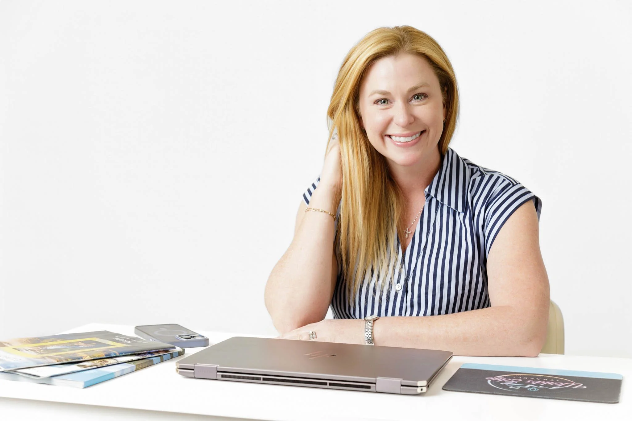 branding photo of smiling woman with strawberry blond hair sitting at white desk with laptop phone and marketing materials white backdrop in studio